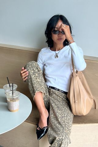 A woman sitting in a cafe wearing a white t-shirt with a black belt, leopard jeans, black pointed kitten heels, and a slouchy tan bag.