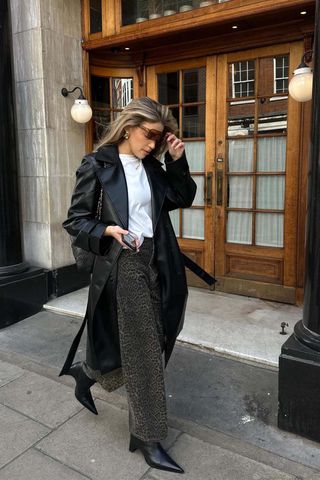 A woman walking down the sidewalk wearing a black leather trench coat over a white t-shirt, dark wash animal print pants, and black pointed ankle boots.