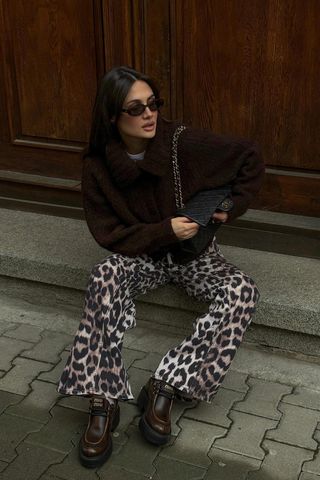 A woman sitting on the sidewalk wearing a white t-shirt under a brown cardigan with leopard pants and brown chunky Miu Miu loafers.