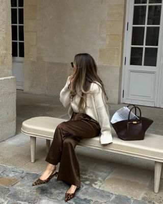 @oliviafaeh wearing a cream sweater, brown silk pants, leopard-print slingback pumps, and a brown Demellier New York Tote