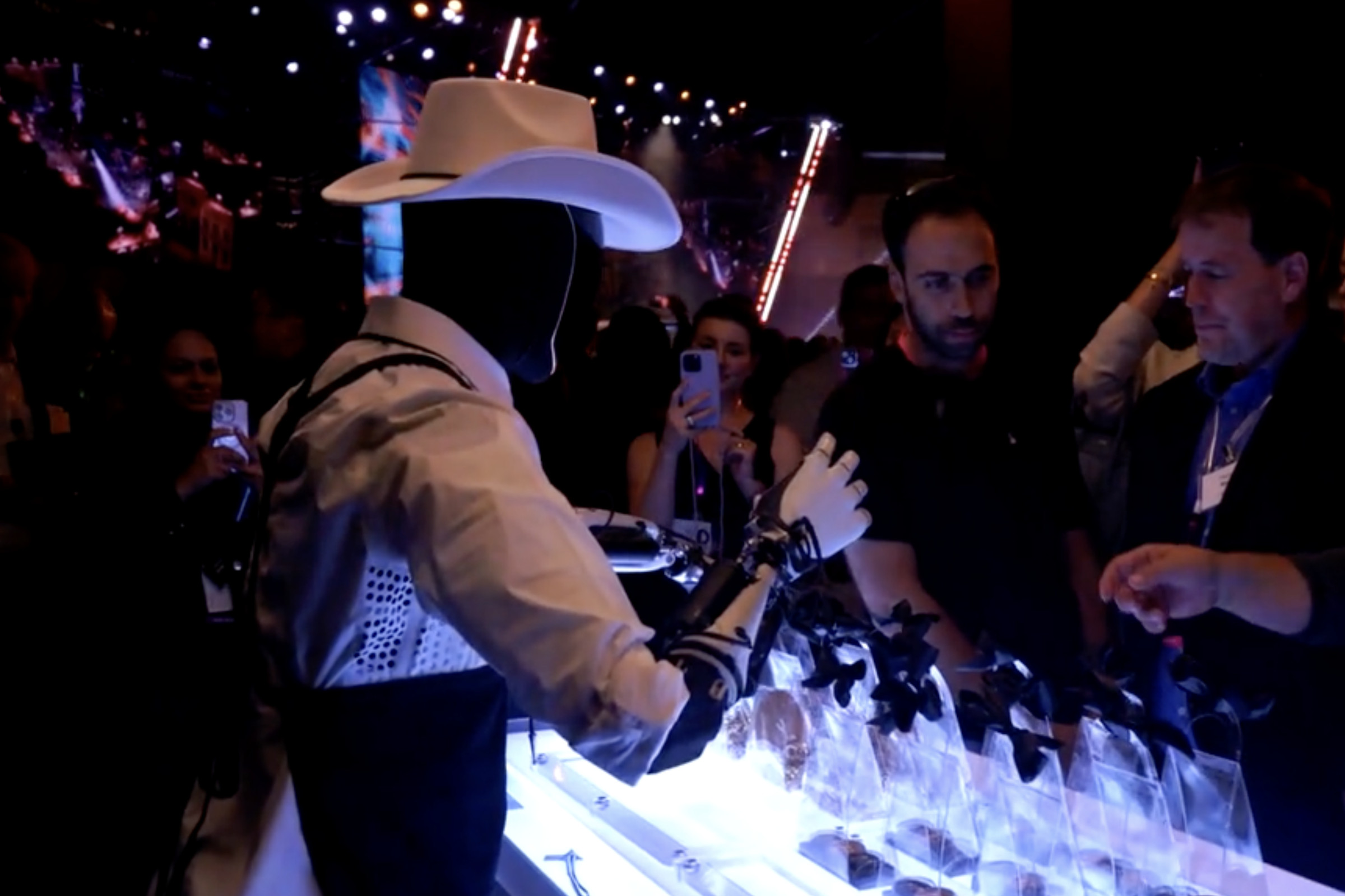 A picture of an Optimus robot, wearing a cowboy hat and standing in front of a crowd of people, behind a table.