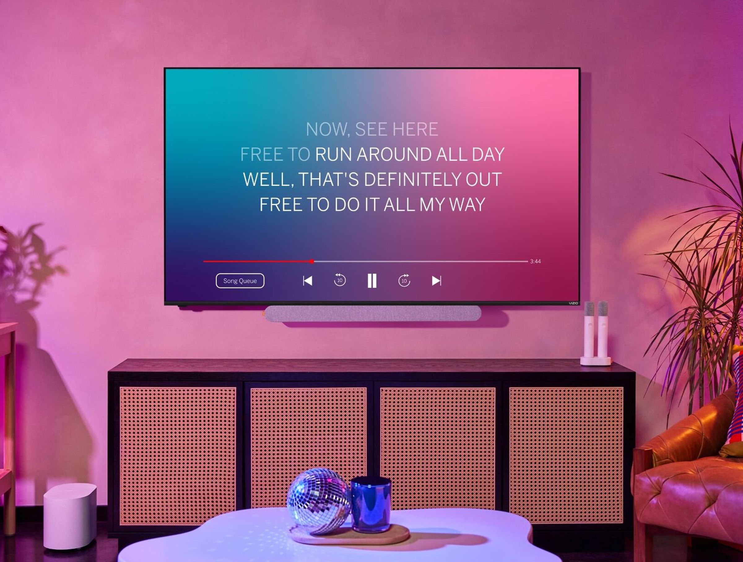 Image of a TV with the MicMe soundbar, two microphones in the charging cradle on a TV console underneath. An image on the screen shows a karaoke song in progress.