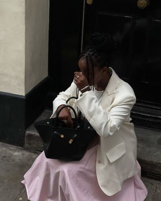 Woman wears a pale pink skirt with a cream blazer and a black bag