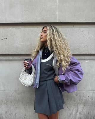 Woman wears a purple sporty jacket with a grey pleated mini skirt and white Miu Miu bag