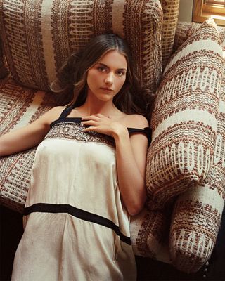 Actress Chloe East lays on a couch wearing a cream dress with embroidered detailing.