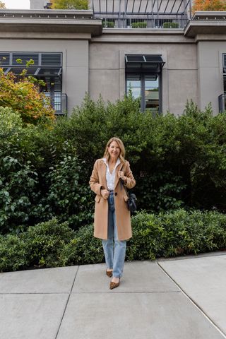 Sandy Koszarek wearing a tan overcoat, white button-down, and light-wash jeans.