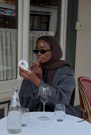 a photo of a woman sitting outside at a table wearing brown sunglasses with a brown scarf over the head with a gray coat looking into a small compact mirror while putting on lipstick