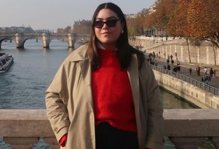 Woman wearing red sweater, classic khaki trench coat, black sunglasses, standing on Paris bridge.