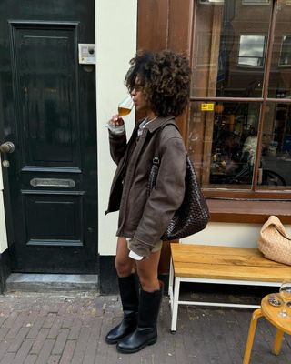 Woman stands outside a cafe wearing black pull on boots and a brown leather jacket