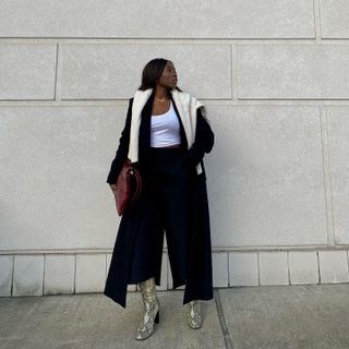 Woman stands in front of a pale wall wearing black trousers, a black coat, and snake print boots