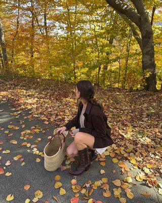 Woman wears brown cowboy boots with a brown jacket and basket bag on a leafy road in autumn