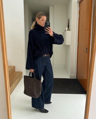 Woman stands in her hallway wearing a navy jumper, blue jeans and square-toe boots