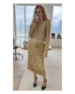 Olympia wears a gold sweater, gold fringe midi skirt, and black mary jane pumps.