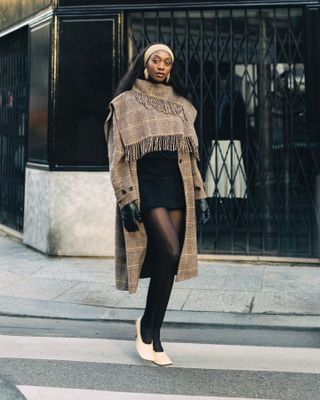 Emmanuelle wears a plaid scarf coat, a beige headband, black minidress, black leather gloves, and ivory pumps.