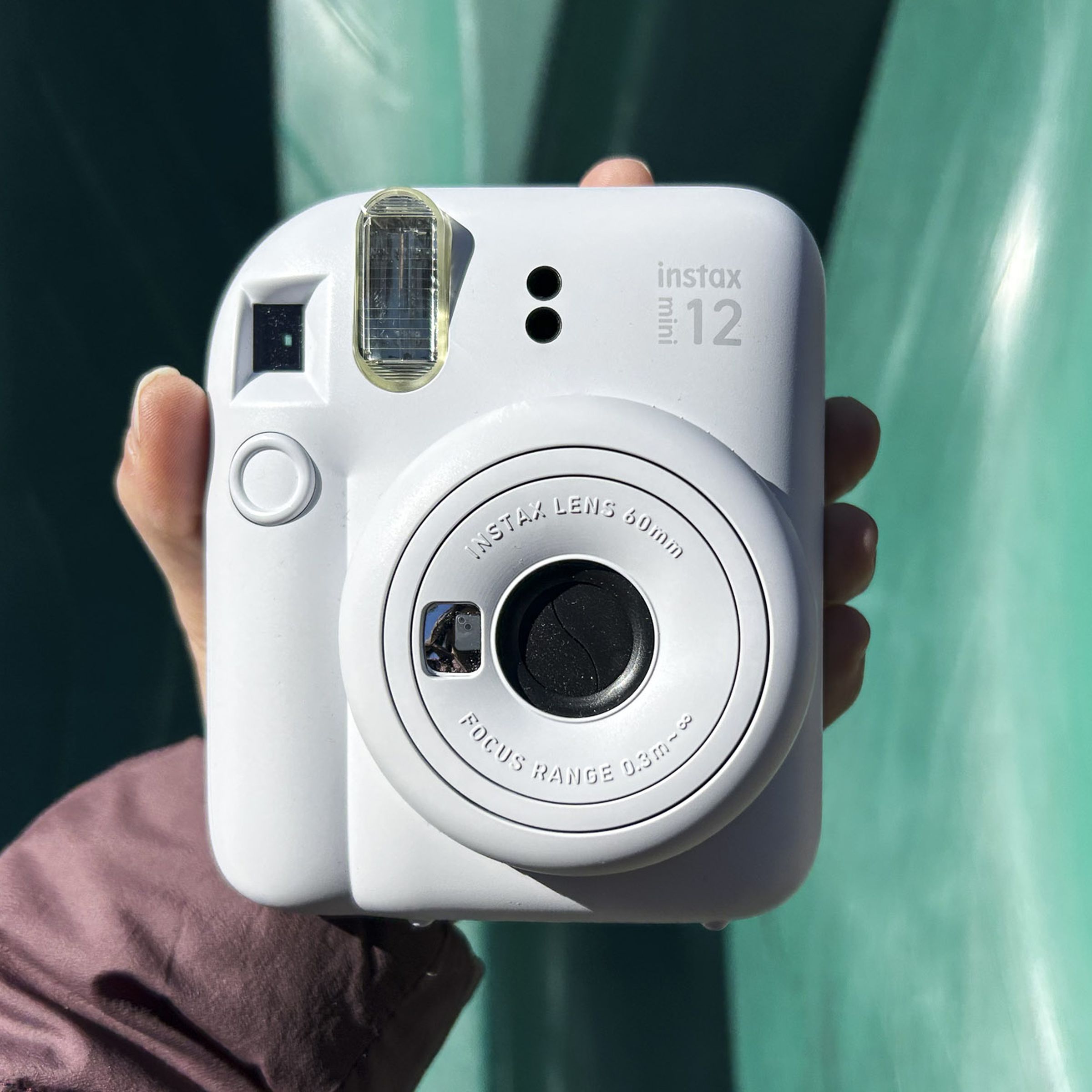 A hand holding up the white Fujifilm Instax Mini 12 against a green slide.