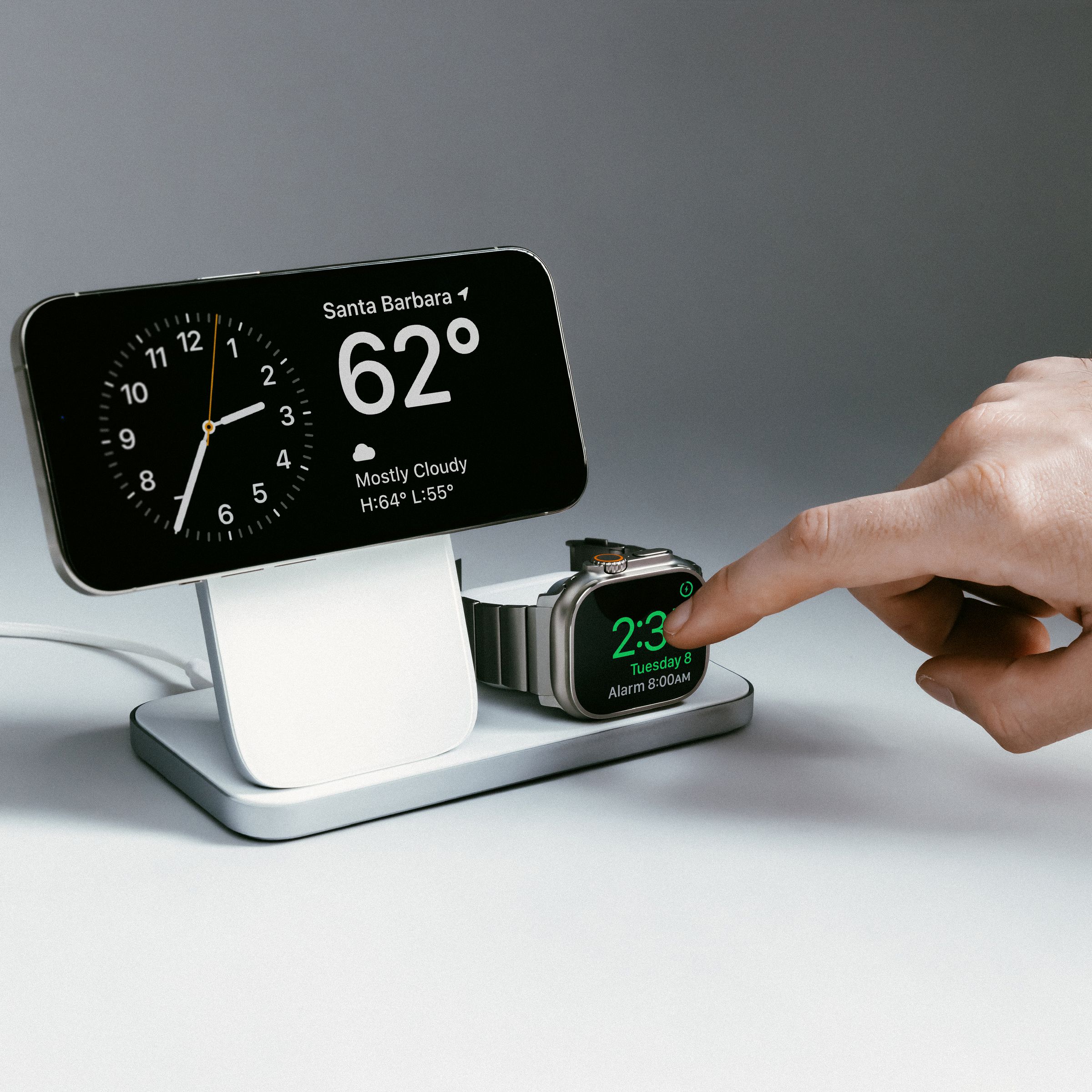 An iPhone and Apple Watch wirelessly charging on a Nomad Stand One Max three-in-one charger. The iPhone is horizontal in StandBy mode and a person is tapping on the Apple Watch screen.