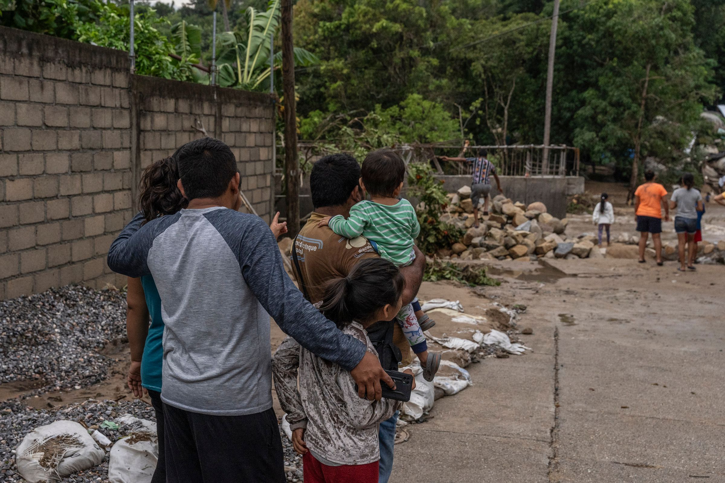 A group of 3 adults and two children are seen from behind, looking out at rubble left behind after a hurricane.