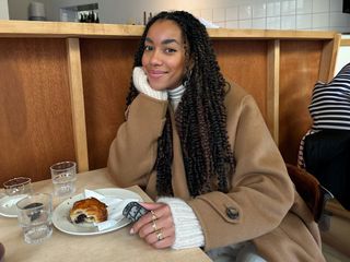 Dutch female fashion influencer Amaka Hamelijnck poses at a cafe wearing a camel coat layered over a chunky white sweater