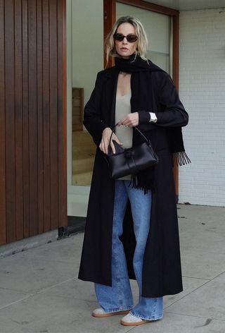 One of the best chunky cuff bracelets is shown in a photo of a woman wearing a long black coat with a silver cuff on the sleeve styled with a tan sweater, jeans, and tan sneakers