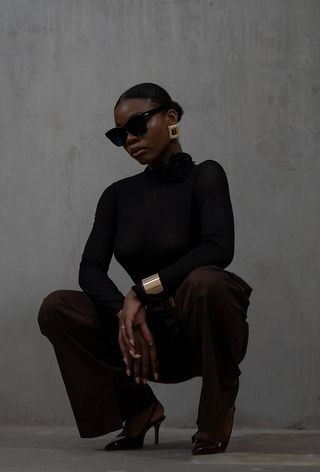 one of the best chunky cuff bracelets shown in a photo of a woman crouching outside wearing black sunglasses, gold earrings, a black turtleneck, black trousers, black pumps, and a chunky gold bracelet