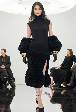 one of the best chunky cuff bracelets shown in a photo from Alaïa's fall 2024 runway show with a model wearing a black turtleneck dress with black leather gloves styled with large gold cuffs and black ankle strap pumps