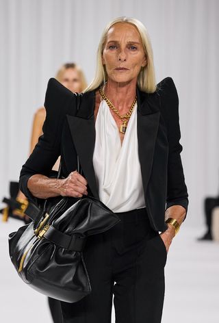 one of the best chunky cuff bracelets shown in a photo from Balmain's spring 2025 runway show with a model wearing a black blazer with dramatic pointed shoulders styled with a white cowl-neck blouse, gold chain necklace, black tote bag, black trousers, and a chunky gold bracelet