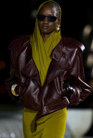 One of the best chunky cuff bracelets is shown in a photo from Saint Laurent's spring 2023 runway show with a model wearing a green hooded jumpsuit under a merlot red leather moto jacket styled with gold clip-op earrings, black sunglasses, and an oversized chunky cuff bracelet