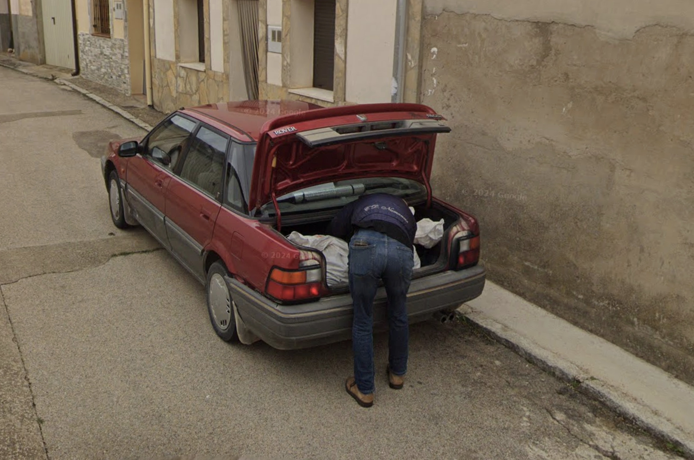 A screenshot from Google Street View showing a man bending over putting something into the trunk of a red car.