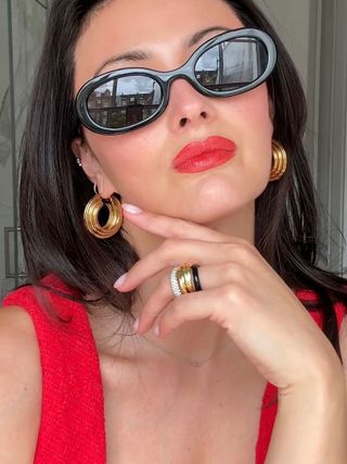 Woman wearing black oval sunglasses, red lipstick, and gold earrings and rings.