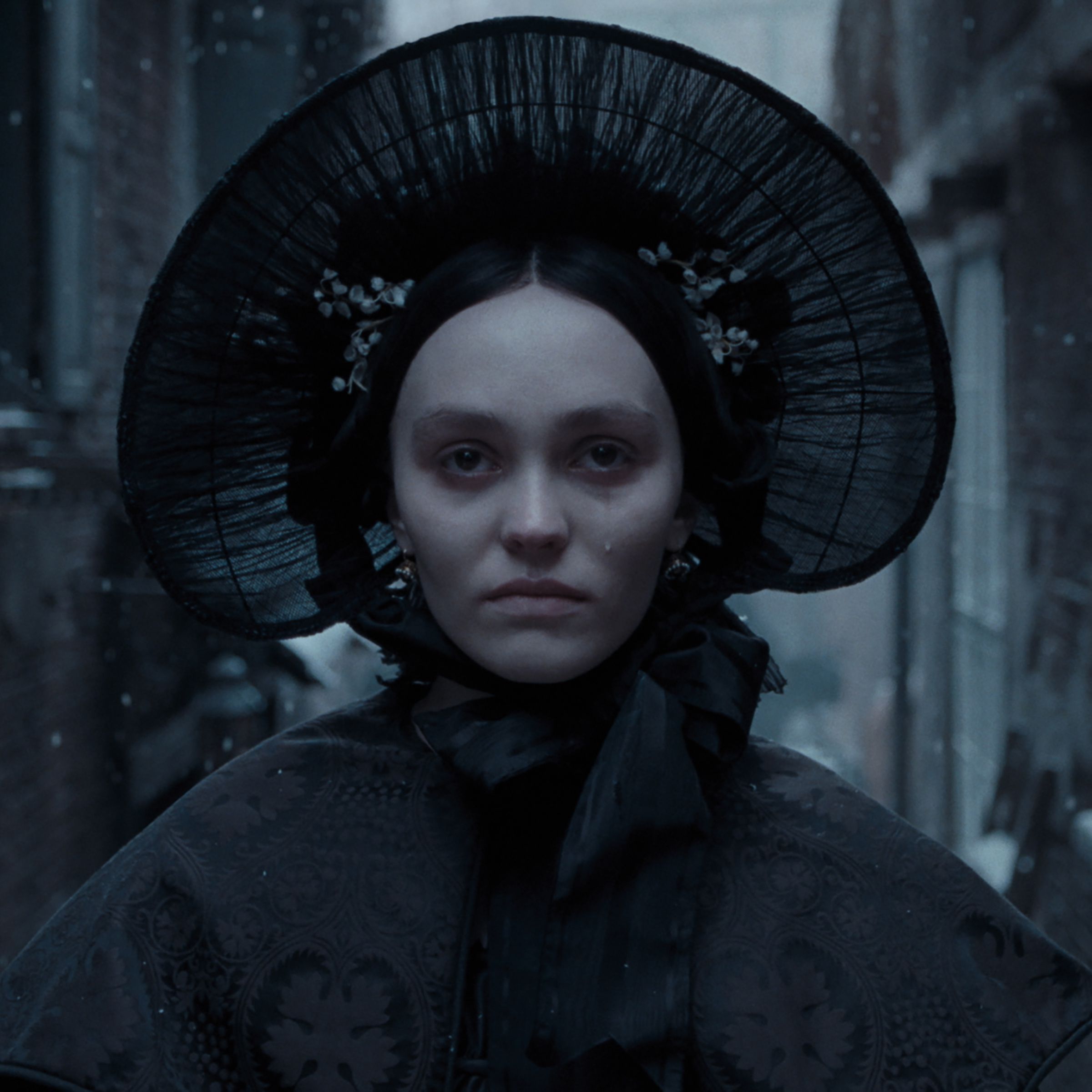 A woman in a black bonnet and black coat standing in an alleyway in winter time.