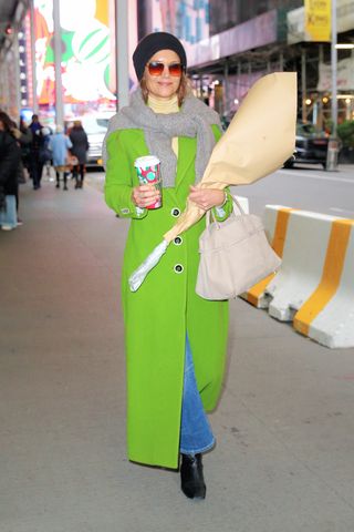 Katie Holmes wears bright green coat, gray scarf, turtleneck, blue jeans, black boots, a black beanie, Manu Atelier bag, and a bouquet of flowers in New York City.