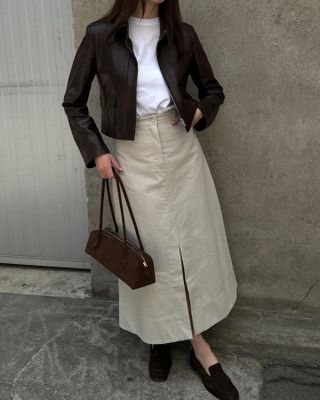 Annabel wears a short black jacket, white t-shirt, khaki skirt, black flats, and a brown suede alaia teckel bag.