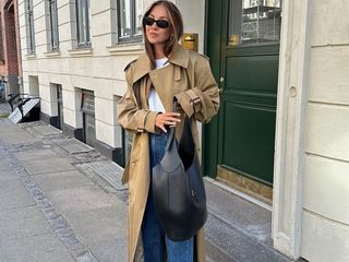 fashion influencer Felicia Akerstrom poses on a sidewalk in Copenhagen wearing black oval sunglasses, a trench coat, white t-shirt, jeans, and black leather shoulder bag