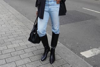 A woman wears a red baseball cap, black blazer, white turtleneck, blue jeans, black knee-high boots, and a black tote bag.