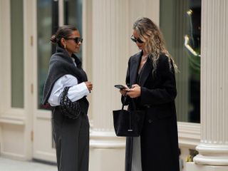 Anouk Yve looking at her phone as she and her friend stand together on a New York street wearing chic, neutral fall outfits.