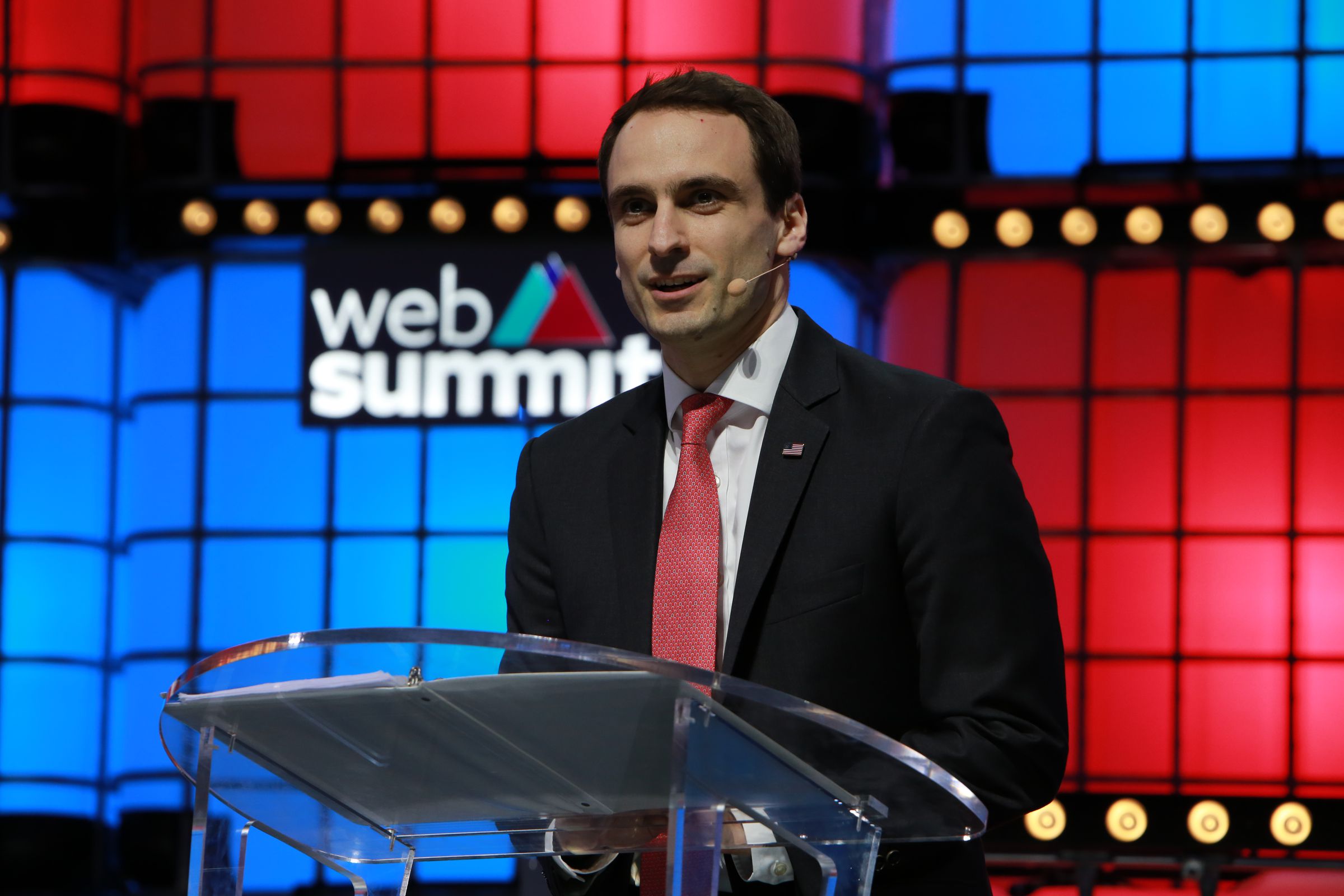 Kratsios standing at a podium with the Web Summit logo on a colorful wall behind him.