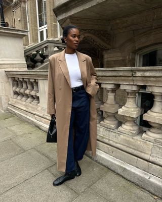 Woman wearing white t-shirt, dark wash jeans, khaki coats, and black flat shoes while standing on London street.