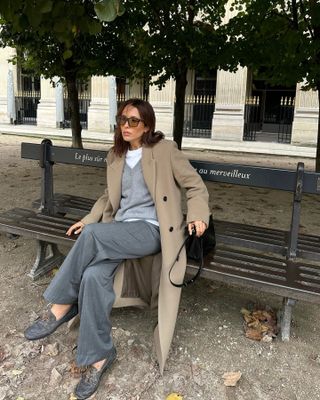 Woman wearing a gray sweater, gray pants, a khaki coat, and boat shoes while sitting on a bench.