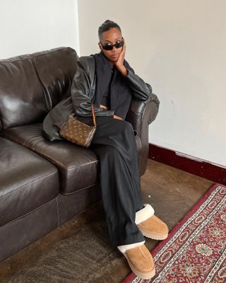 Woman wearing leather jacket, black pants, and platform shearling Ugg slippers while sitting on a couch.