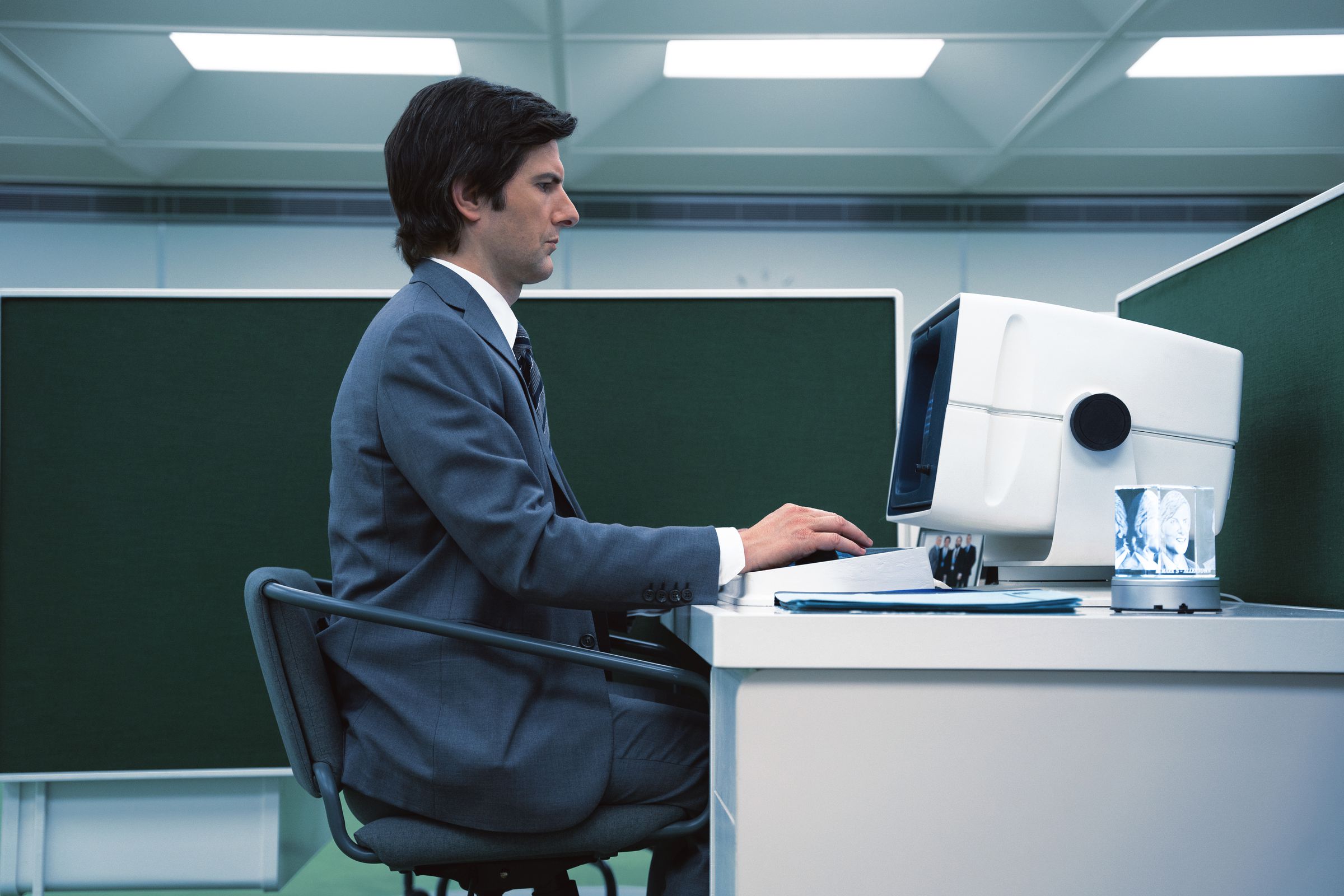 A man in a gray suit sitting at his desk and typing on a computer in a cubicle.