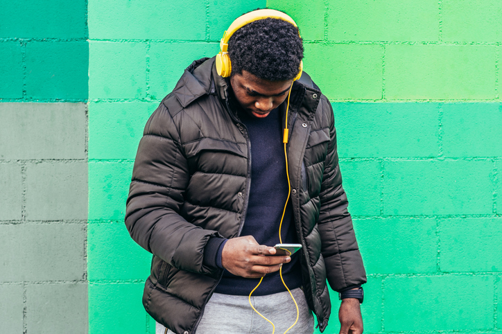 Black African American boy on green background wall. Listening to music with yellow headphones and his mobile phone.