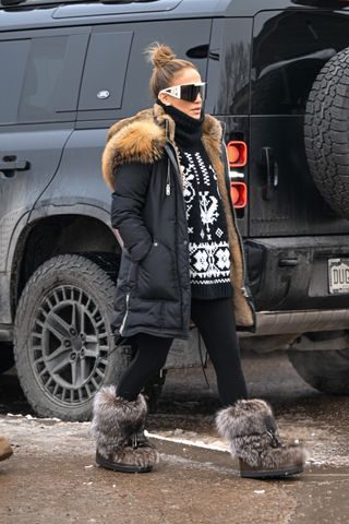 Jennifer lopez wears large sunglasses, a black turtleneck sweater, black fur-lined winter coat, and black leggings.