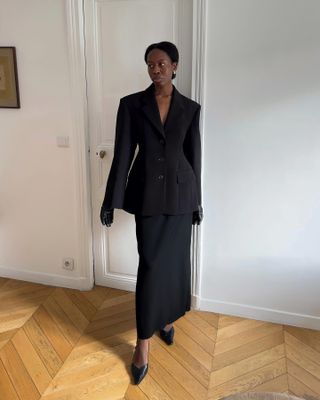 Sylvie wears an oversize black blazer, black maxi skirt, black leather gloves, and black heels.