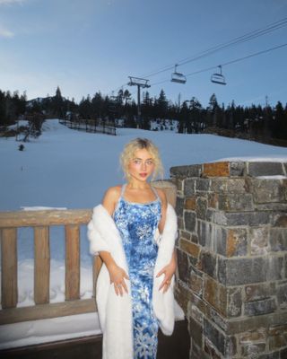 Sabrina Carpenter posing in front of a ski lift wearing a blue snowflake dress and fairy dust eye shadow.