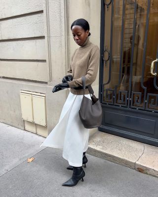 Sylvia wears a tan sweater, white skirt, brown leather bag, black heels, and black leather gloves.